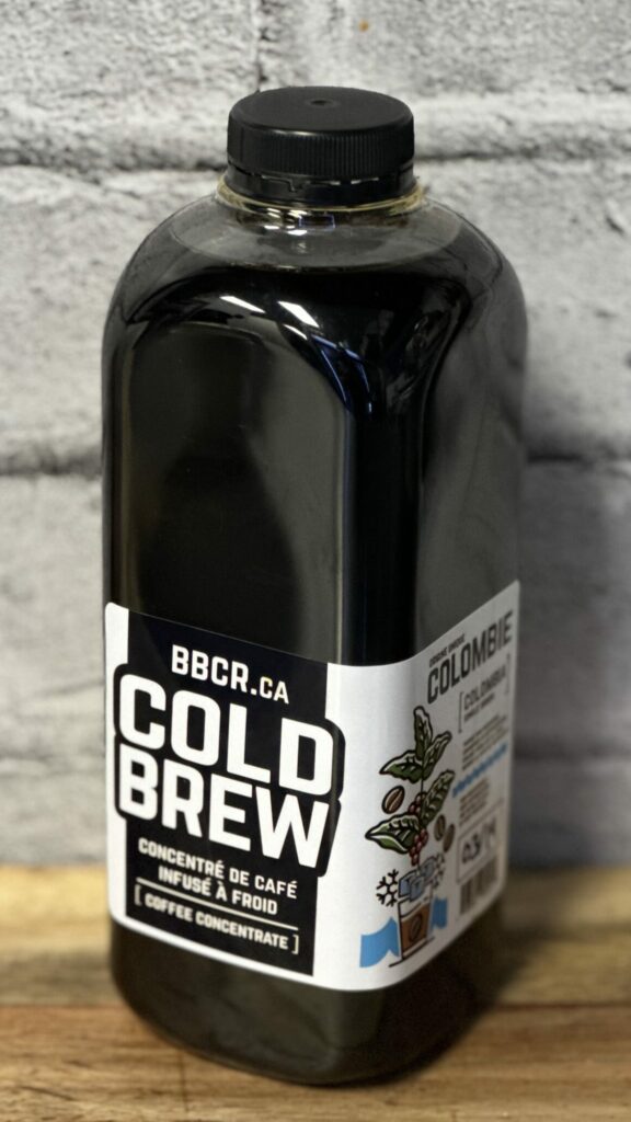 Bottle of cold brew coffee concentrate labeled BBCR.ca, standing on a wooden surface against a gray brick wall.